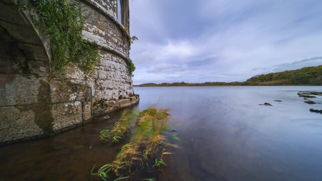 Timelapse of a medieval tower on lake with shoreline rocks and reeds in the foreground and forest in the distance on a cloudy day in county Roscommon in Ireland.