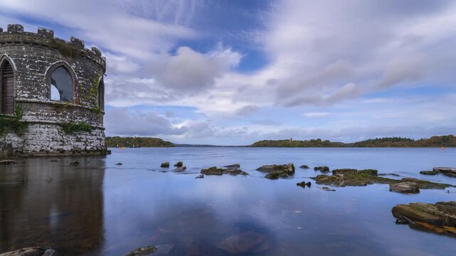 Timelapse of a medieval tower on lake with shoreline rocks in the foreground and medieval castle in the distance on a partially cloudy day in county Roscommon in Ireland.