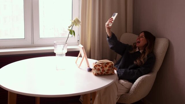 A young woman sits in a grey armchair by a window, holding her smartphone up to take a selfie. She is smiling and showing a "V" or peace sign with her hand. The scene includes a white table with a mir