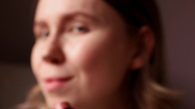 A young woman shows a brown contouring cosmetic stick to the camera and then applies the cream product to her cheekbone while smiling joyfully. Natural makeup and face sculpting concept