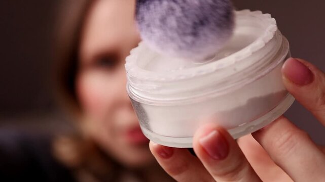 A young woman performs a beauty routine by dipping a pink brush into a jar of translucent powder and playfully blowing a cloud of product toward the viewer