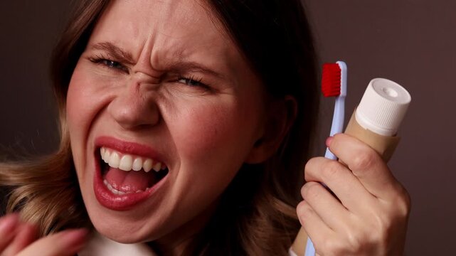 A close-up portrait of a young woman with a joyful, ecstatic facial expression and open mouth, holding a blue toothbrush and a tube of toothpaste against a dark background