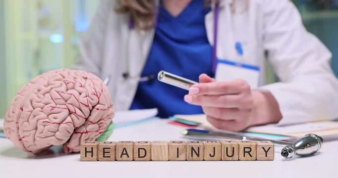 Wooden cubes form phrase Head Injury near anatomical model of brain. Doctor holds diagnostic flashlight preparing to assess neurological responses