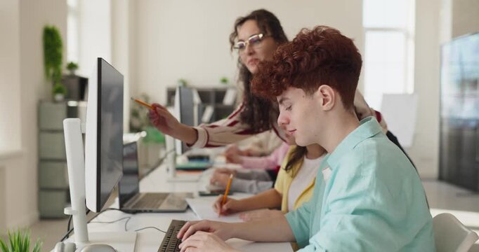 Students and teacher in classroom coding lesson. Focused teenagers code at computers as instructor guides. Modern lab supports collaboration. Concept highlights digital education and teamwork.