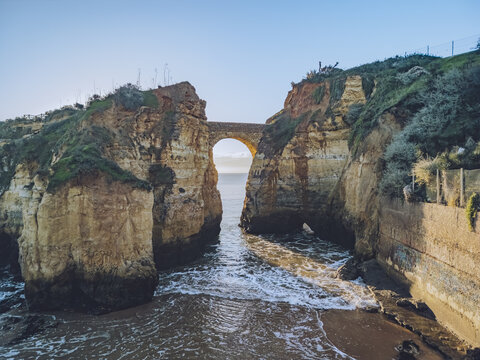 Aerial view of a stone bridge arching over rugged cliffs and crashing waves, sunlight painting the scene with golden hues, Lagos, Faro District, Portugal.