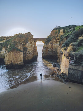 Aerial view of cliffs framing a hidden beach and bridge, where the soft sand meets the rough rock, creating a tranquil coastal scene, Lagos, Faro District, Portugal.