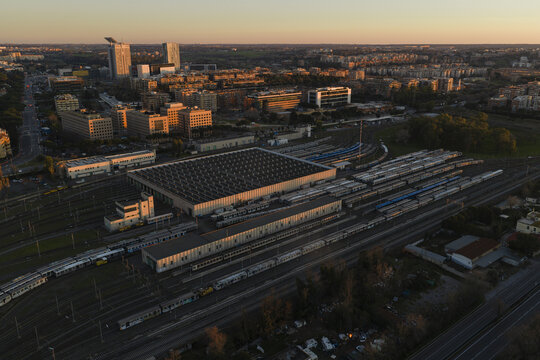 Aerial view of a sprawling train yard bathed in the warm glow of the setting sun, contrasting with the cool tones of the city skyline, Roma, Lazio, Italy.