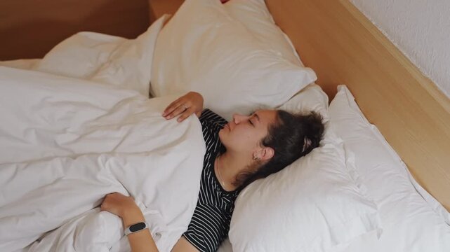 Female traveler napping in hotel bed, relaxed limbs under white duvet, arms overhead and subtle turning, polkadot top, bedside wood headboard, quiet restful pause during trip recovery