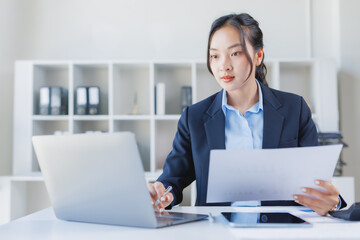 Fototapeta premium Professional businesswoman working in a modern office, analyzing financial reports and documents while typing on a laptop, demonstrating dedication and efficiency in her corporate role