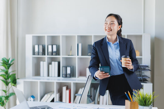 Asian businesswoman standing in a modern office, smiling confidently while holding a disposable coffee cup and using a smartphone, showing daily work and communication