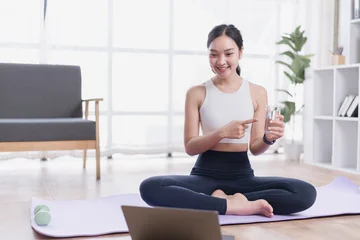 Fotobehang Lotusbloem Young woman sitting in lotus position on a yoga mat with a laptop, pointing at a glass of water while participating in an online fitness class at home  © MINAE