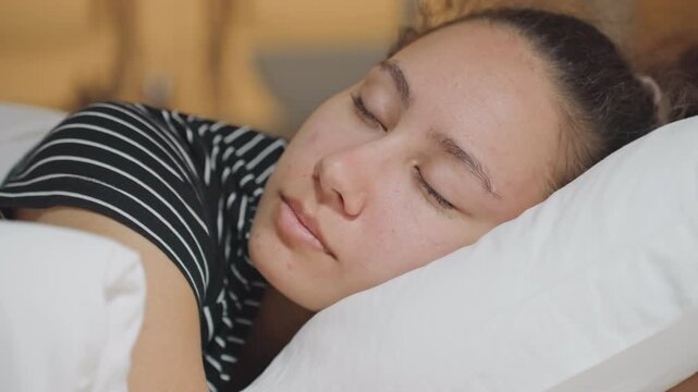 Calm multiracial female resting on bedding with tranquil expression in luxury hotel. Tranquil multiracial woman lies peacefully on pillow in elegant hotel setting with warm lighting