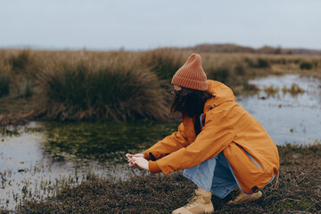 Woman in orange jacket and beanie squats near a pond in a natural landscape with grass and water....