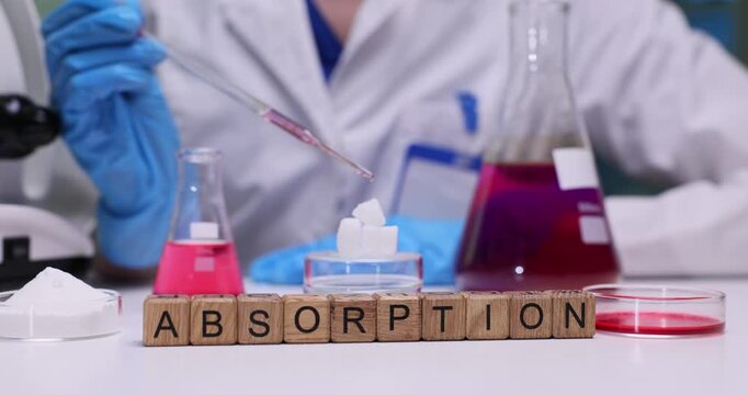 Laboratory worker uses pipette to apply liquid reagent onto white cube-shaped material. Miniature wooden cubes on table form word Absorption