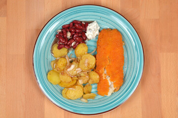 A top down view of a breaded fish fillet served with potato salad and red kidney beans on a blue ceramic plate over a wooden table