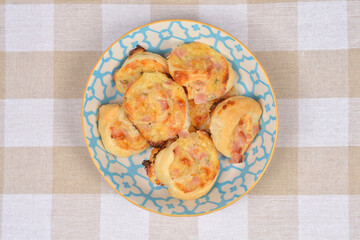 Golden baked puff pastry rolls with ham and cream cheese served in a blue patterned bowl on a checkered tablecloth background