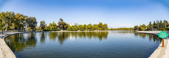 Naklejka premium Panoramic view of a calm lake with tree reflections in Sorgun Titreyengol. Scenic landscape with green foliage, blue sky, and hotel buildings in Antalya province, Turkey. Sunny autumn day.