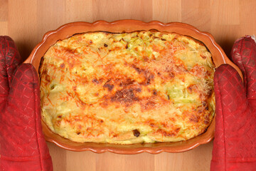 Top view of a freshly baked golden leek gratin in an oval ceramic baking dish held by red oven mitts on a wooden table