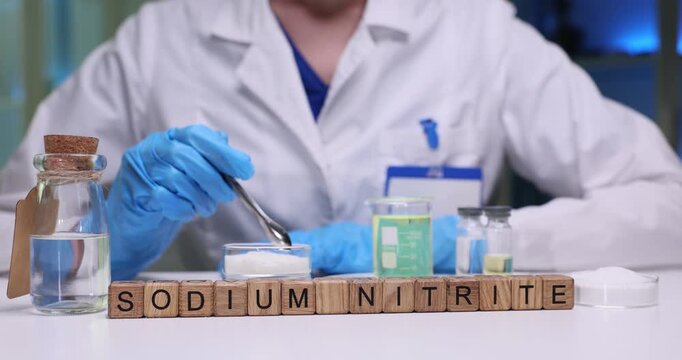 Laboratory worker carefully observes fine texture of white powder inside Petri dish. Wooden cubes form substance name Sodium Nitrite on table