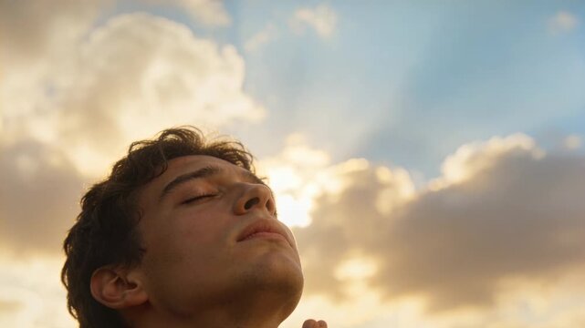 Praying male in collared shirt tilting head under partly cloudy sky, holding clasped hands
