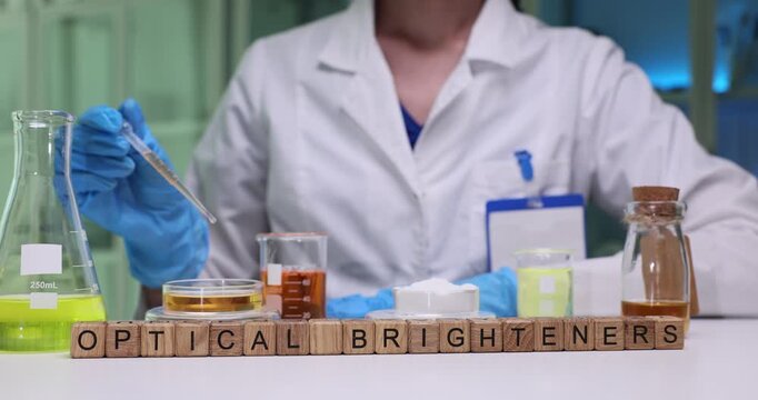 Laboratory worker carefully transfers liquid reagent into Petri dish with fluid using pipette. Miniature wooden cubes form phrase Optical Brighteners