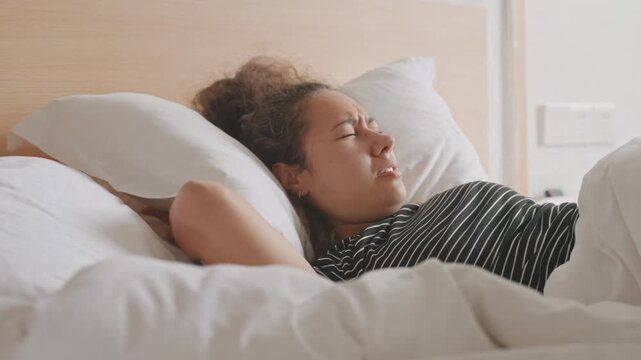 Hotel tourist covering face with pillow on soft white duvet, wooden headboard framing scene slow breaths and hidden expression convey deep exhaustion after long travel day and attempt to block out