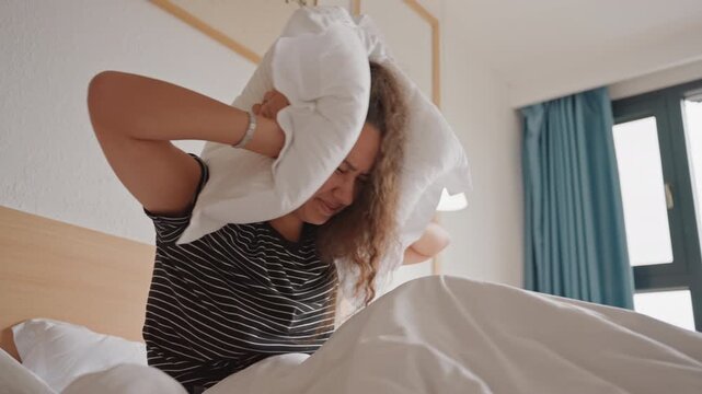 Caucasian woman wakes groggy hotel bed muffling noise with pillow sits at bed edge squinting toward window messy duvet teal curtains pendant lamp frame modern cozy hotel interior morning frustration