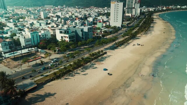 Da nang city bayfront featuring a long sandy beach along the urban coastline, busy street with moving vehicles, and numerous buildings at the foot of distant mountains, aerial view