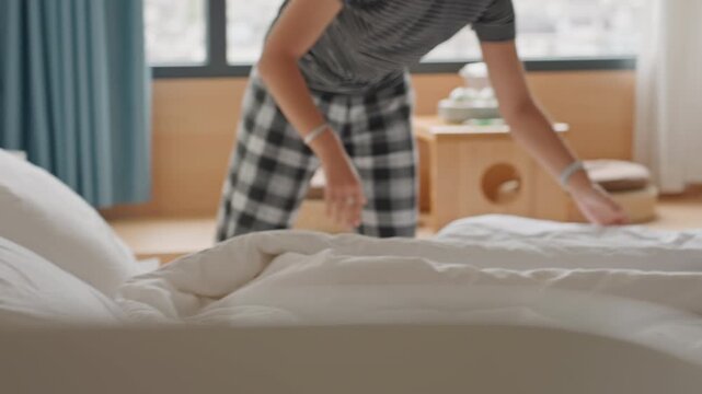 Hotel staff fluffing pillow with care in cozy hotel room, plaid pants and patterned top, bedside lamp and window view, smoothing pillowcase and aligning duvet, preparing room for guest arrival,