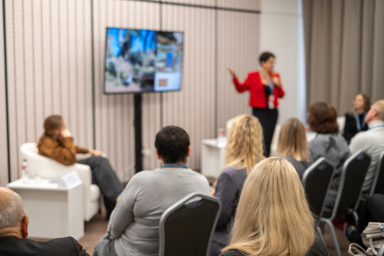 Business conference presentation, speaker addressing seated audience in small professional meeting room, focused discussion and networking atmosphere.