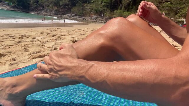 Man applying sun lotion on his tanned leg while relaxing at sandy tropical beach on a hot, sunny day. Skin care, sunblock protection, summer vacation and seaside lifestyle concepts