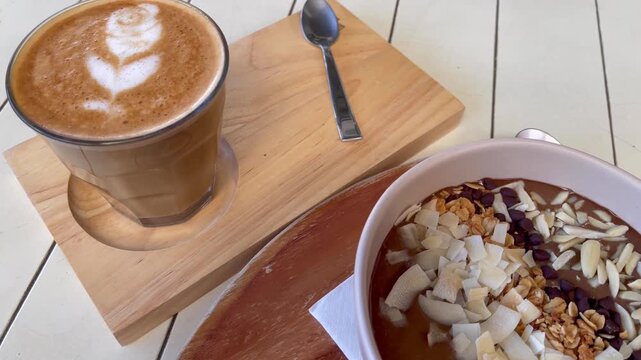 Coffee with heart-shaped latte art served alongside a chocolate smoothie bowl topped with granola, coconut flakes, nuts, and chocolate chips. Cozy cafe breakfast or brunch setting on a rustic table