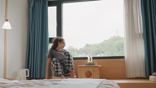 Guest in hotel room morning young woman moves to window sits on bench sets cup on wooden table gazes at green skyline beyond soft natural light through blue curtains bedside lamp neatly made bed calm