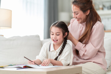 Joyful Asian mother smiles while braiding her young daughter's hair as the little girl happily focuses on painting in her coloring book in a bright modern living room.
