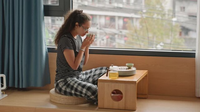 Woman in hotel room watching city, small table with tea and journal, sunlight filtering through curtains, thoughtful posture, planning itinerary, quiet urban stay energizing reflective mood