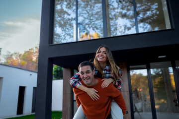 Fototapeta premium Cheerful couple embracing new home ownership by piggybacking and smiling against a modern house exterior, symbolizing new beginnings, success, and a happy future together