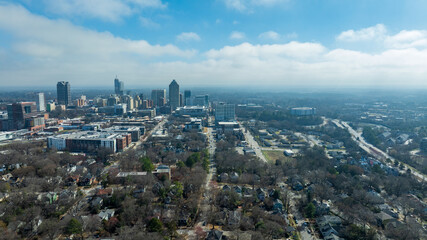 Naklejka premium Downtown Raleigh skyline with highway, residential neighborhoods, trees, and urban landscape under bright cloudy sky.