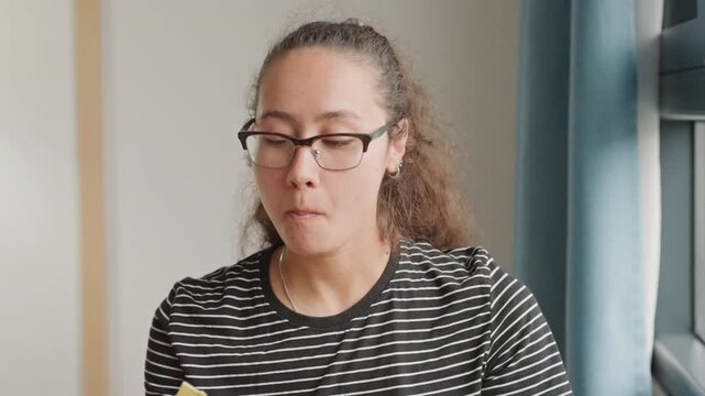 Multiracial woman guest hotel window calm morning scene sips tea bites cookie while gazing outside striped shirt glasses soft daylight blue curtains porcelain cup reflective mood solo business