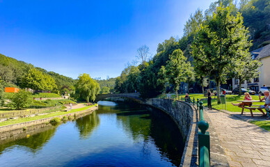 Fototapeta premium Esch-sur-Sure, Wiltz, Grand-Duche de Luxembourg, September 07, 2025, sunny river scene with stone bridge and riverside seating, cheerful blue sky and people-friendly promenade where local artist