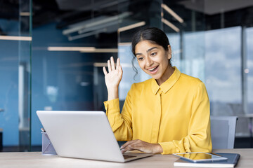 A young Indian woman sits in the office at a desk in front of a laptop, talking on a video call, smiling and greeting the camera