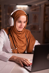 Female Muslim doctor in hijab and white coat using headphones and laptop computer during...