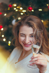 beautiful young woman winks and holds a glass of champagne on the background of a New Year tree