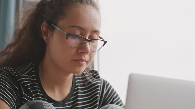 Young woman working laptop hotel window seat, focused expression, glasses and striped shirt, editing documents and scrolling notes, quiet morning light, remote worker balancing planning and fatigue
