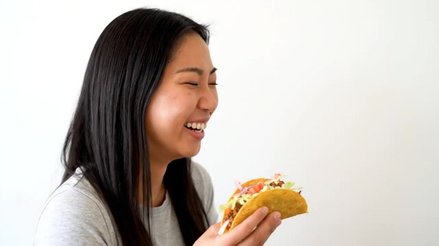 A young woman with long black hair, wearing a gray shirt, is eating a taco, initially smiling broadly, then gradually showing different expressions of amusement and contemplation, against a plain bac