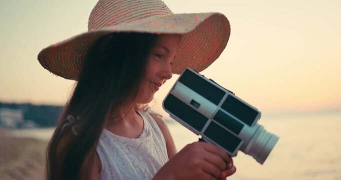 Little girl with hat, dress and old vintage camera making photos and videos of sea waves and beach, seascape sunrise. Young photographer.