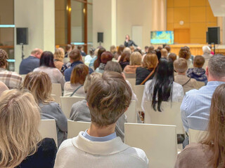 Fototapeta premium Audience seated in hall listening to speaker, business conference with screen. Education Workshop Session. Forum Presentation. Professional Networking Meeting. Corporate Training Seminar, training