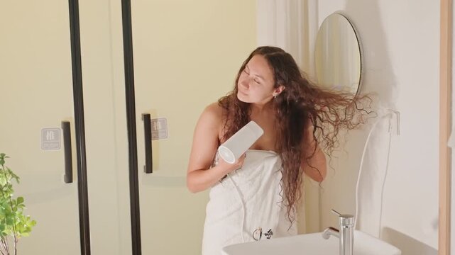 Hotel bathroom guest relaxing hair, towel wrapped romantic suite vibe with soft lamp light and round mirror, gentle hairdrying and playful smile, intimate honeymoon stay and elegant decor