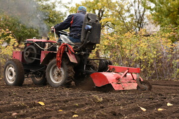 Obraz premium Man on red tractor plowing the field, side-back view, smoke from chimney, horizontal shot.