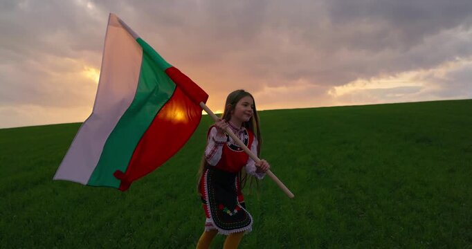 Girl in bulgarian folklore costume and Bulgarian flag running on green field at sunset cinematic video