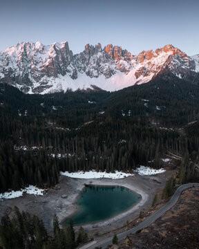 Aerial view of a tranquil lake nestled amidst a dense forest, with snow-capped mountains kissed by the morning light in the background, Carezza, Trentino-South Tyrol, Italy.